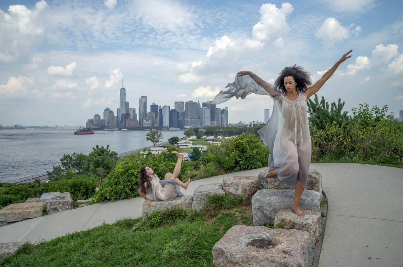 Gabrielle Willis sits on rocks, extending one leg with a flexed foot. Natasha Diamond-Walker stands on a rock, extending her arms in a V. Behind them is the downtown Manhattan.